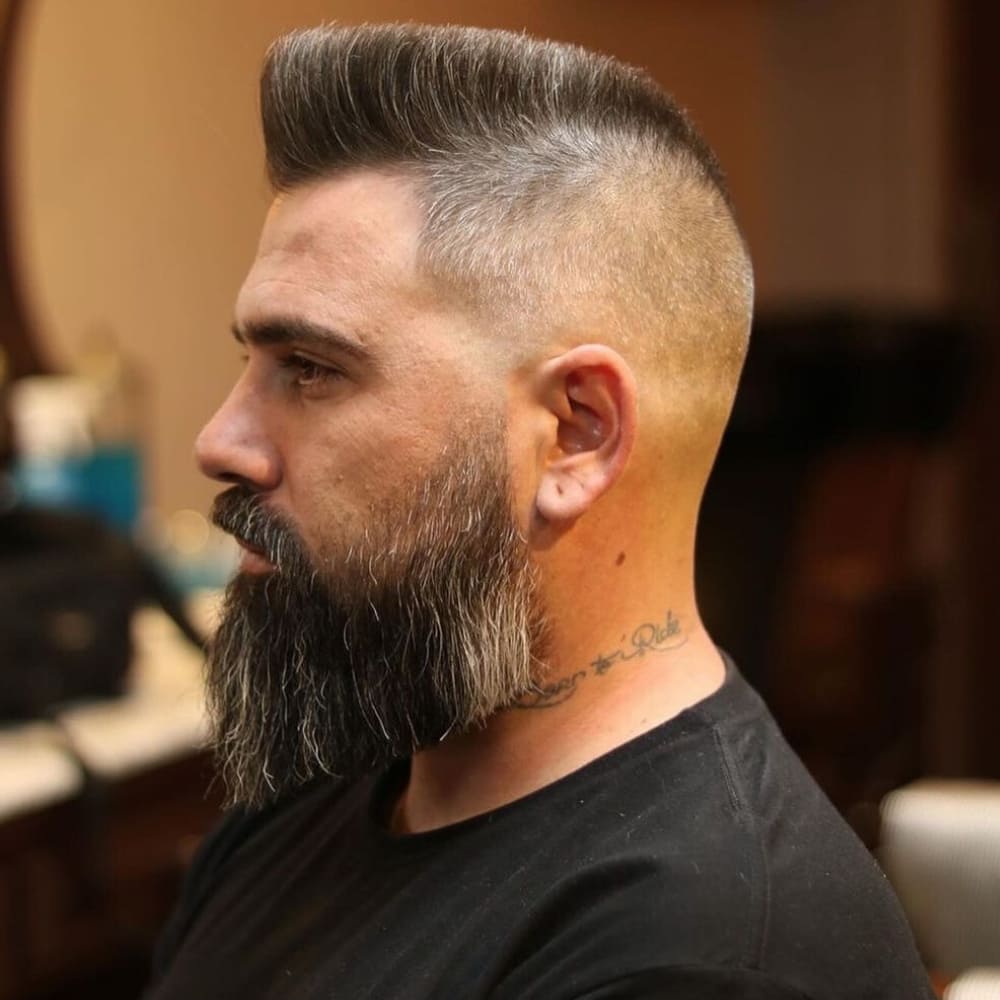 Side profile of a man with a classic flat top, featuring sharply defined edges and a high, flat surface. Paired with a full beard and a clean fade on the sides, this bold haircut exudes confidence and a retro vibe, perfect for those looking to make a strong style statement.