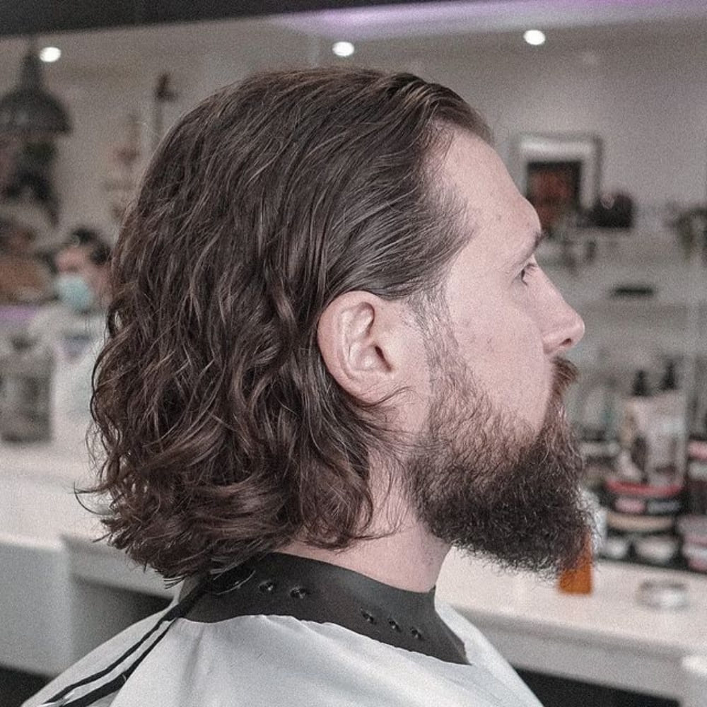 Man with straight hair styled in loose waves, featuring shoulder-length hair with natural texture and gentle waves. He has a full beard and is sitting in a barbershop chair, wearing a barber cape, showcasing a relaxed and effortless look in a modern barbershop setting.