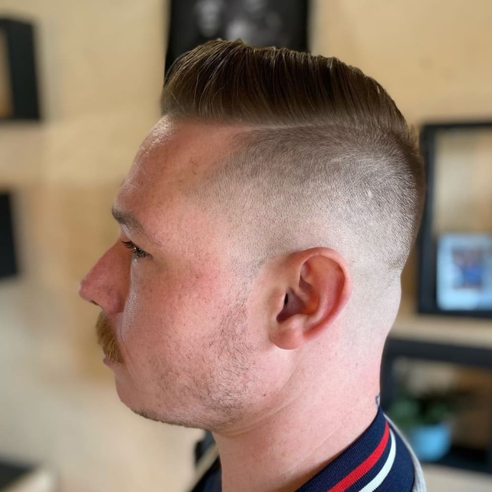 A man with a disconnected pompadour haircut is pictured from the side in this image. His hair on top is styled in a sleek, voluminous pompadour, while the sides are sharply shaved, creating a clear disconnect between the lengths. The neat, precise lines of the shave emphasize the bold contrast. The man's mustache adds a classic touch to the modern haircut. The background of the image is a softly lit indoor space, suggesting a professional barbershop setting. The overall look is clean, stylish, and contemporary.