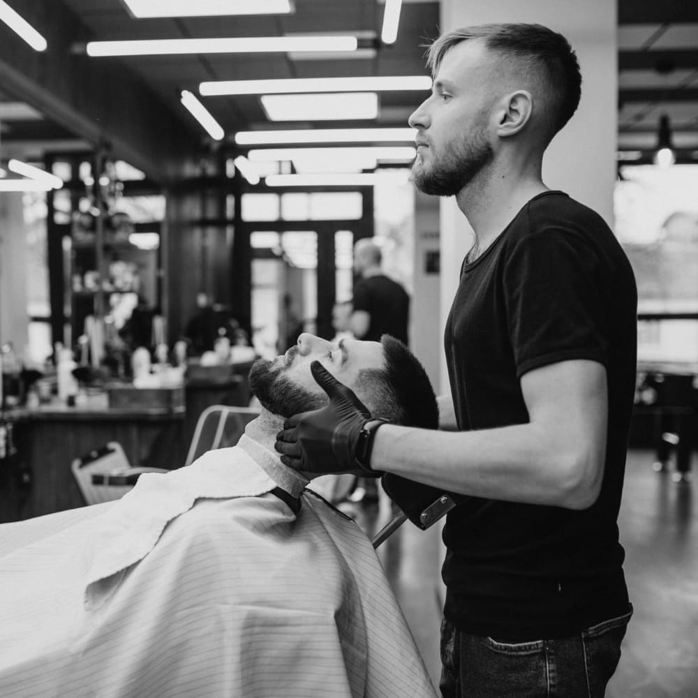 A black and white photograph of a barbershop scene, where a professional barber with a neatly trimmed beard and a short haircut is giving a shave to a client. The barber, wearing a black t-shirt and gloves, holds a straight razor while gently positioning the client’s head back for precision. The client, draped in a striped barber cape, has a well-groomed beard and a fresh haircut. The background features a modern barbershop interior with mirrors, grooming tools, and other clients receiving haircuts, creating a stylish and professional atmosphere.