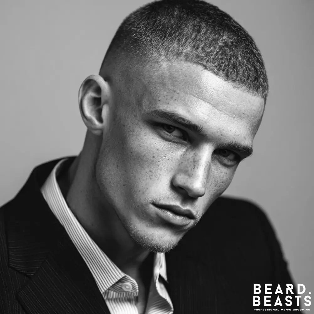 A close-up, black and white photograph of a man with a sharp buzz cut fade. The sides are faded high and tight, blending seamlessly into the short, textured hair on top, showcasing a clean and modern professional men's hairstyle.
