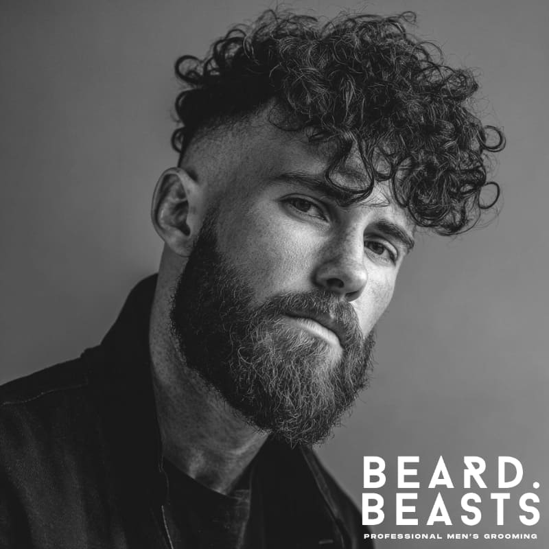 Close-up of a man with a curly hairstyle featuring short faded sides and voluminous curls on top. Paired with a full beard, this short on sides, long on top haircut creates a bold, modern contrast between sharp fades and natural curl texture.