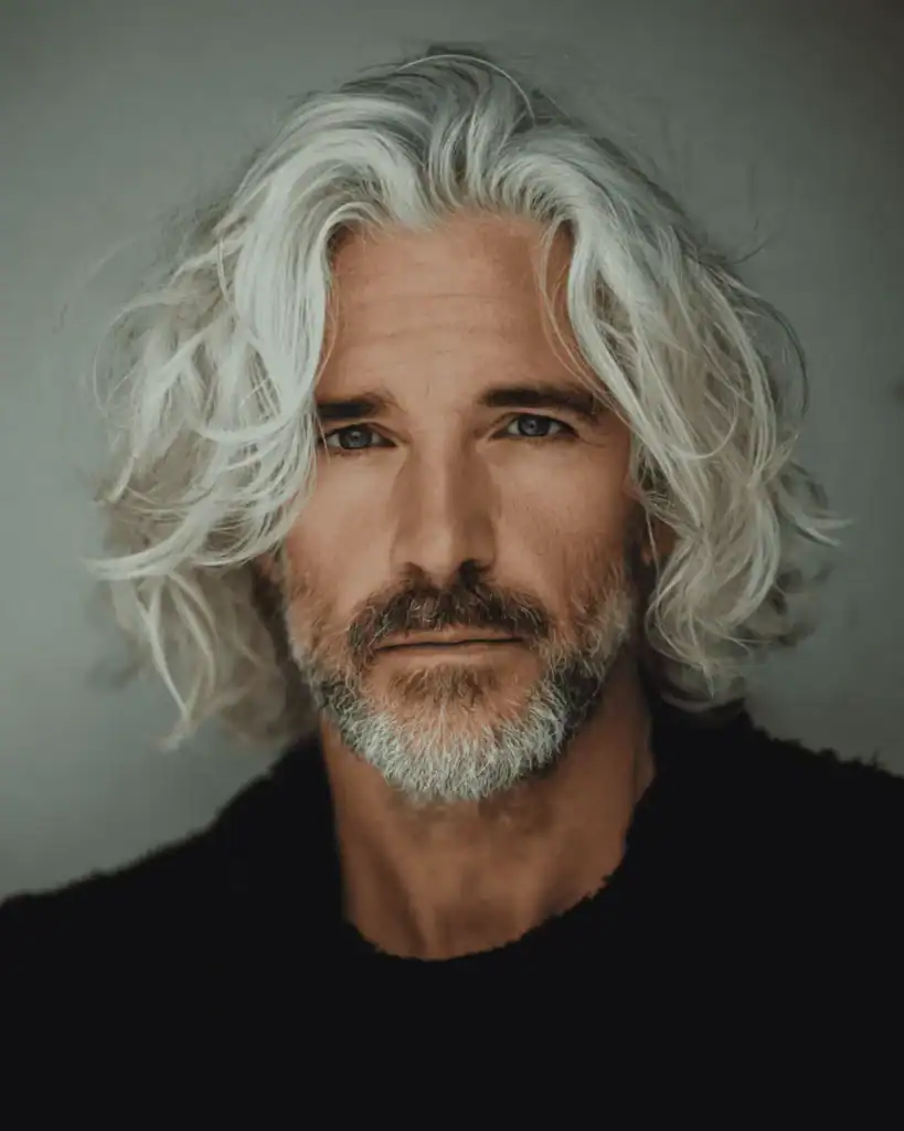 A portrait of a rugged older man with shoulder-length layered silver hair, featuring loose waves that frame the face to soften angular features.