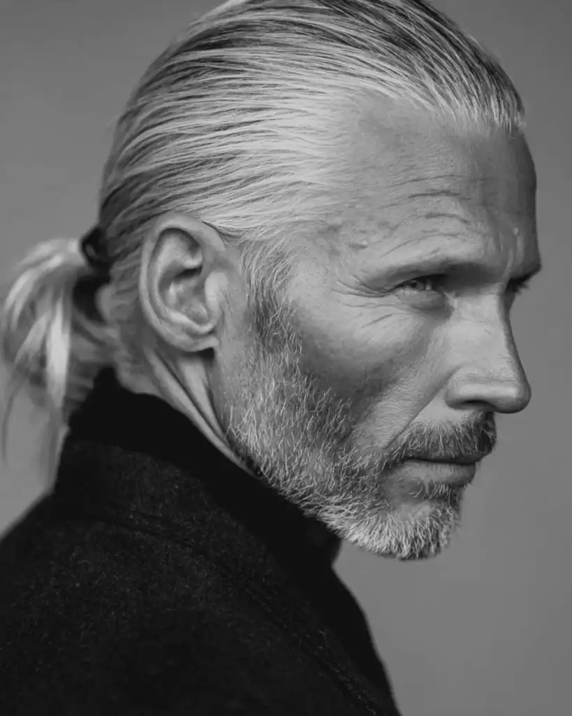 A black and white profile portrait of a distinguished older man with a low ponytail hairstyle, featuring silver hair pulled back and tied securely at the nape of the neck.