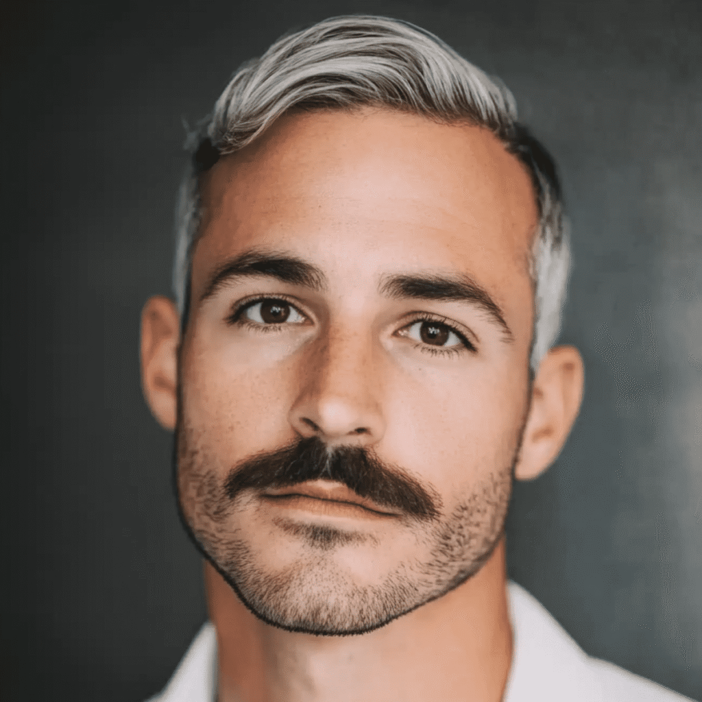 A stylish man with silver-gray hair sports a well-groomed Chevron mustache in this image. His hair is neatly combed to the side, adding a modern touch to his look. The mustache is thick, full, and perfectly shaped, covering his upper lip in the classic Chevron style. His beard is trimmed short, maintaining a clean and polished appearance that complements the mustache. He wears a crisp white shirt, which contrasts beautifully with his tanned skin and silver hair, giving him a fresh, sophisticated look. The background is a dark, blurred gradient, ensuring that the focus remains on his sharp facial features and expressive eyes. The overall image exudes confidence, style, and a modern take on a timeless mustache style.