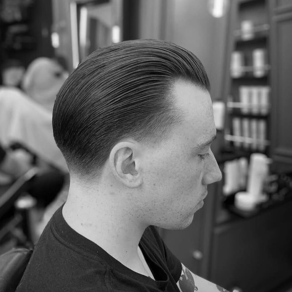 A black and white photograph of a man with a tapered slick back hairstyle, sitting in a barbershop. His hair is neatly combed back with a smooth, glossy finish, likely styled with pomade. The taper fade on the sides transitions cleanly from a shorter length near the ears to a longer top. The background features a blurred barbershop setting with grooming products and another client receiving a haircut. The man has a sharp, structured appearance, emphasizing the polished and professional nature of the tapered slick back style.