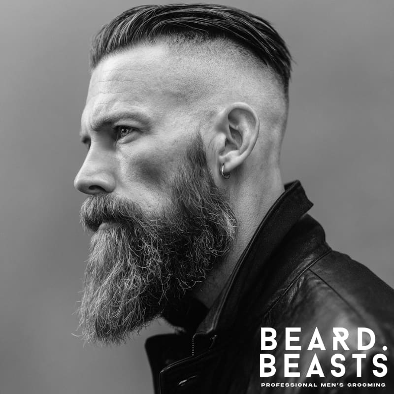 A bold example of men’s short on sides, long on top haircuts, featuring a high skin fade with longer hair on top styled back. The sharp fade on the sides contrasts with the voluminous, slicked-back hair on top, complemented by a full, well-groomed beard. This modern, edgy look offers a clean, structured finish with a touch of rugged style.