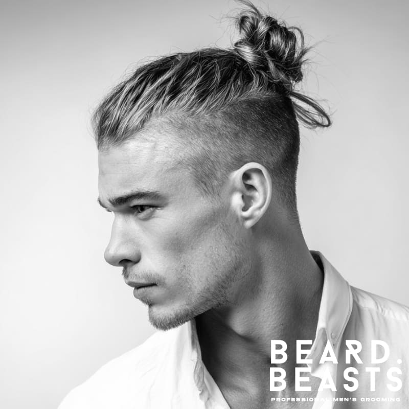 A modern example of men’s short on sides, long on top haircuts with a high skin fade and a stylish man bun on top. The clean fade on the sides contrasts with the longer hair styled into a casual bun, offering a trendy and effortless look with a sharp, well-defined silhouette.