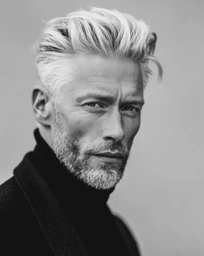 A black and white portrait of a stylish older man with a modern pompadour hairstyle, featuring silver hair styled with significant height and volume swept back from the forehead.