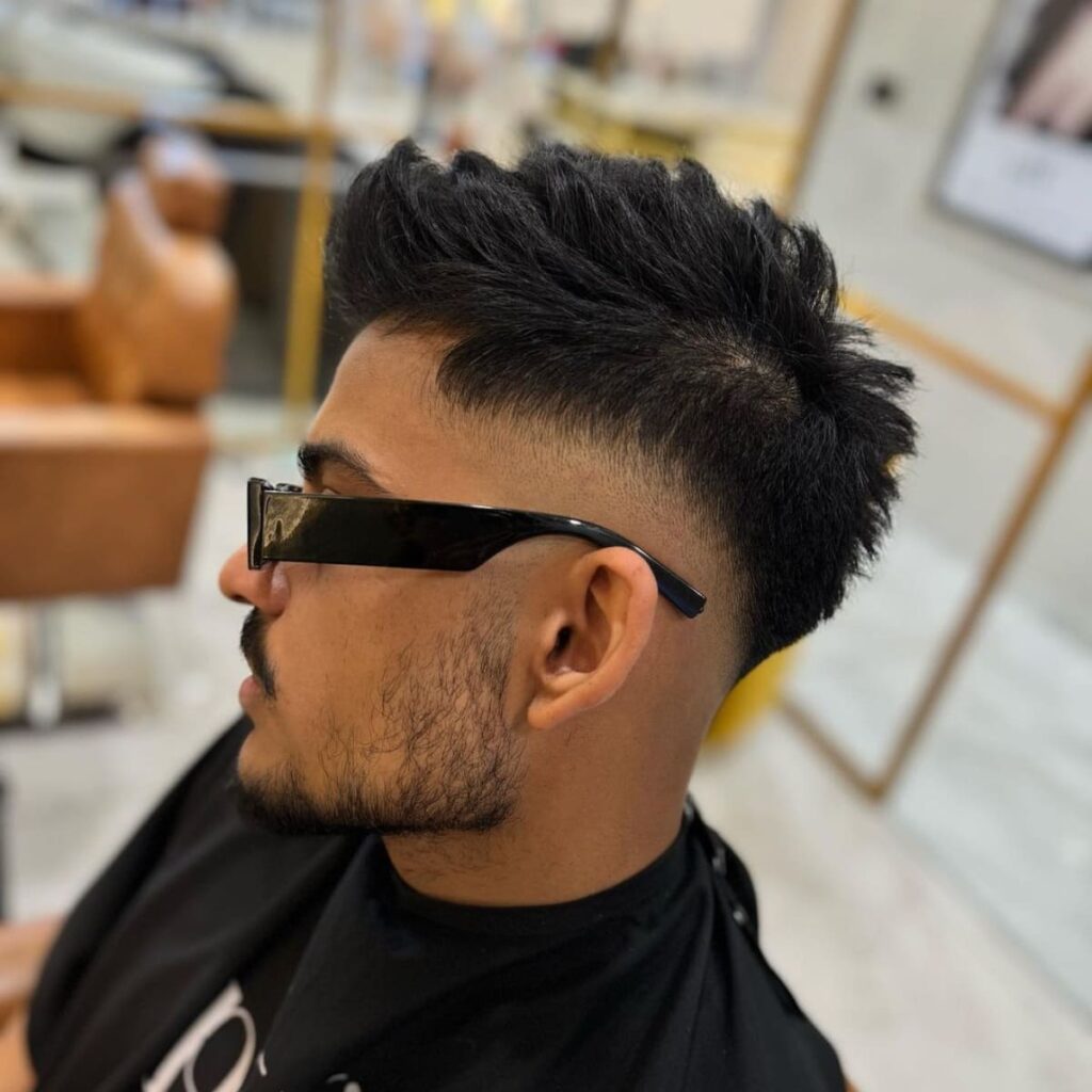 Side profile of a man with a mid haircut, showcasing a smooth transition from a full, textured top to closely shaved sides around the middle of the head. The hairstyle adds volume on top, styled upward for a dynamic, modern look, while the taper enhances the clean lines and contrasts sharply with the longer hair. Paired with stylish sunglasses, this look offers a trendy, confident vibe.