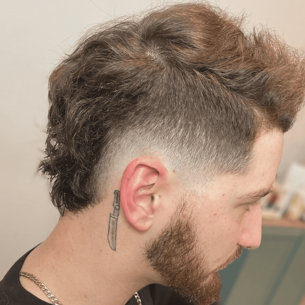 Side profile of a man with a low fade haircut, textured hair on top, and a gradual fade around the ear. The style is complemented by a beard and a knife tattoo behind the ear, creating a distinctive and edgy appearance.