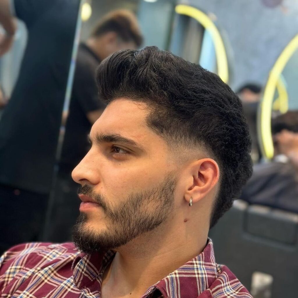 Side profile of a low burst fade quiff hairstyle featuring a rounded fade around the ears and a high-volume, textured top. This low burst fade quiff is ideal for adding a modern, edgy look with natural waves and a well-groomed beard for added style.