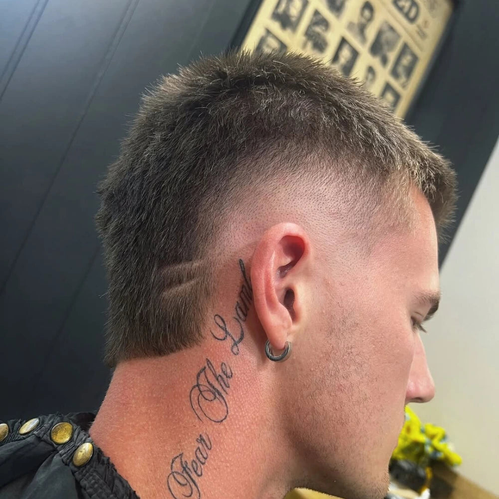 Man with a textured burst fade buzz cut and two shaved hairline designs behind the ear, paired with a neck tattoo and hoop earring.