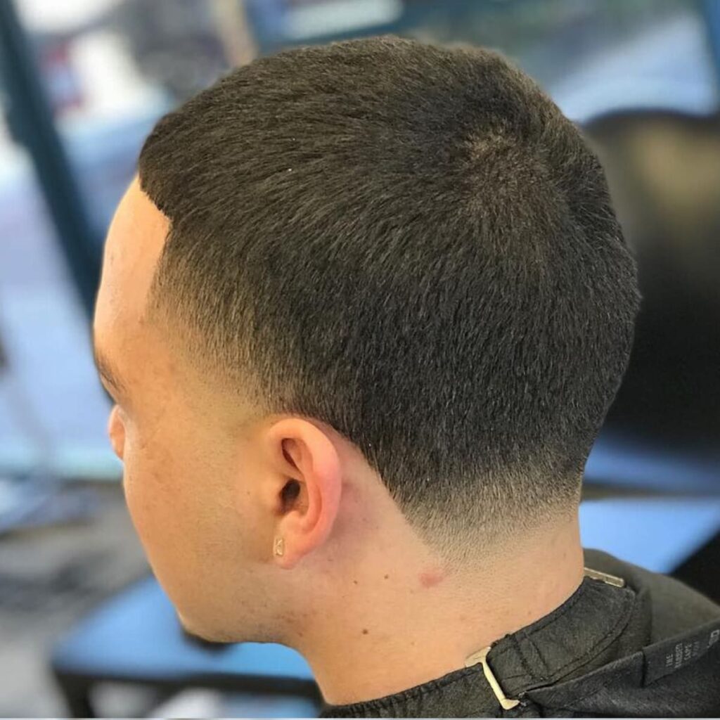 A back and side view of a man with a skin fade buzz cut, showcasing a gradual fade that transitions cleanly from the skin at the neckline to short, even hair on top. The haircut is precise, with sharp edges and a polished, textured finish. This low-maintenance style combines a sleek fade with simplicity, making it both modern and versatile.