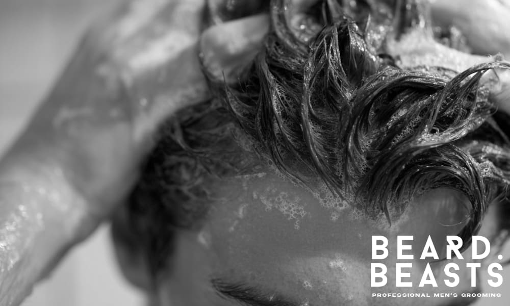 Close-up of a man washing his hair, with visible shampoo lather in the strands. The image captures a healthy haircare routine in a natural setting.