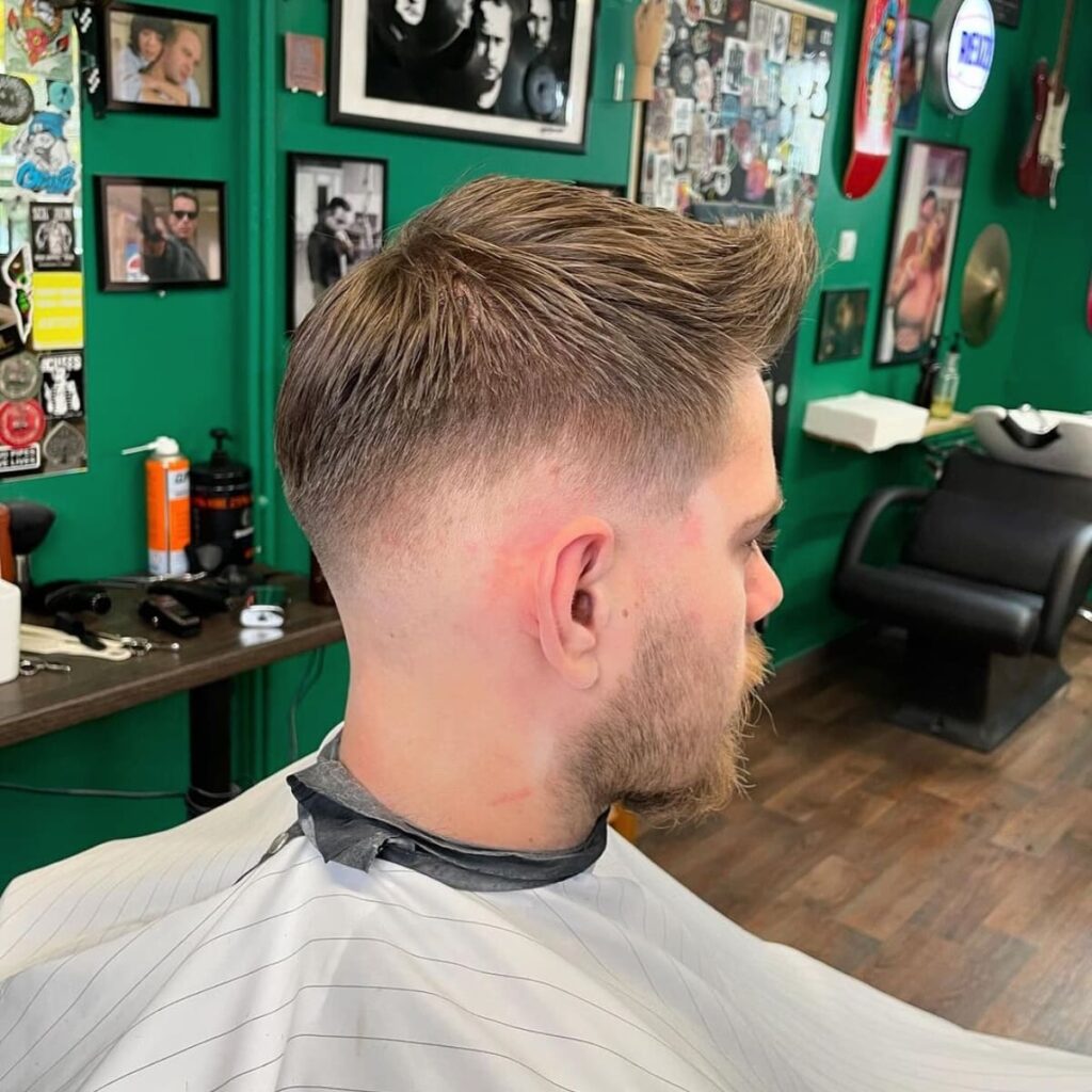A man sits in a barbershop chair, showcasing a fresh haircut with a voluminous top styled upward and slightly back. The sides are cleanly faded, creating a sharp and seamless transition. He has a light beard that complements the hairstyle. The barbershop features a vibrant green wall adorned with framed photos, artwork, and guitar decor, creating a lively and eclectic atmosphere. The workspace is organized with grooming tools and products visible in the background.