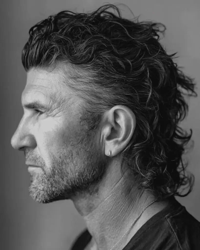 Black and white profile of a man over 40 with a modern textured mullet, featuring curly hair with length at the back and softly tapered sides.