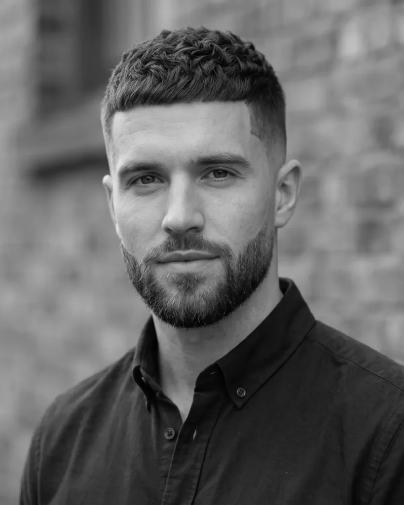 A white man with a short textured top haircut and a clean low fade, featuring a perfectly shaped and trimmed beard with a sharp cheekline against a blurred brick background.