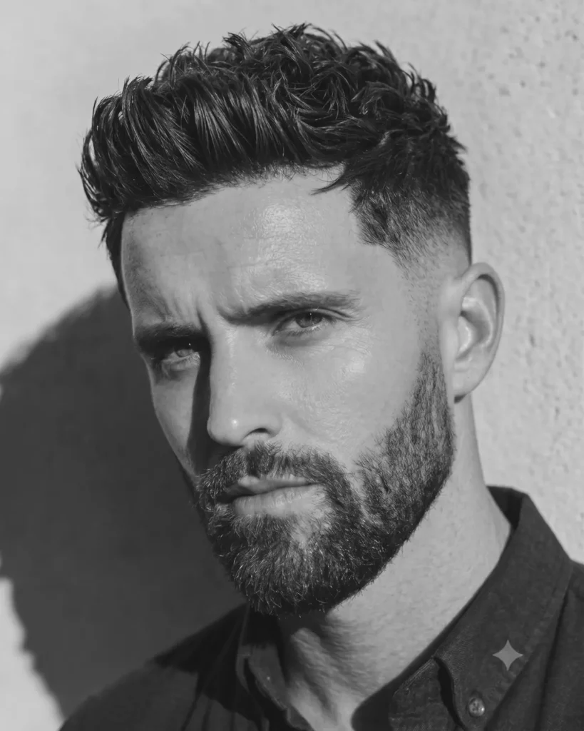 Man with a textured quiff, low fade haircut and full beard in a black and white portrait.