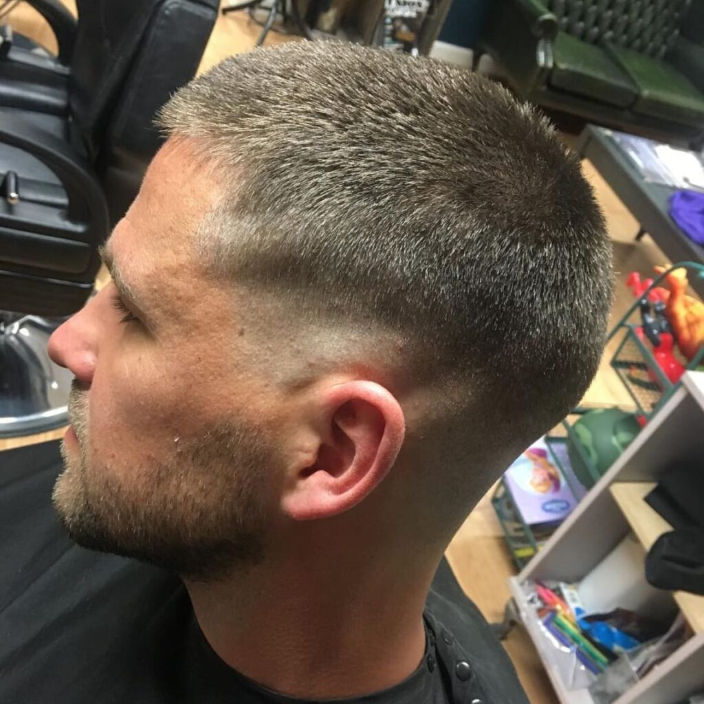 Side profile of a man with a clean and precise haircut featuring closely cropped sides that blend seamlessly into the slightly longer hair on top. The beard is neatly trimmed, complementing the sharp lines of the haircut. The setting is a modern barbershop, with leather chairs and grooming tools visible in the background, creating a professional and polished atmosphere.