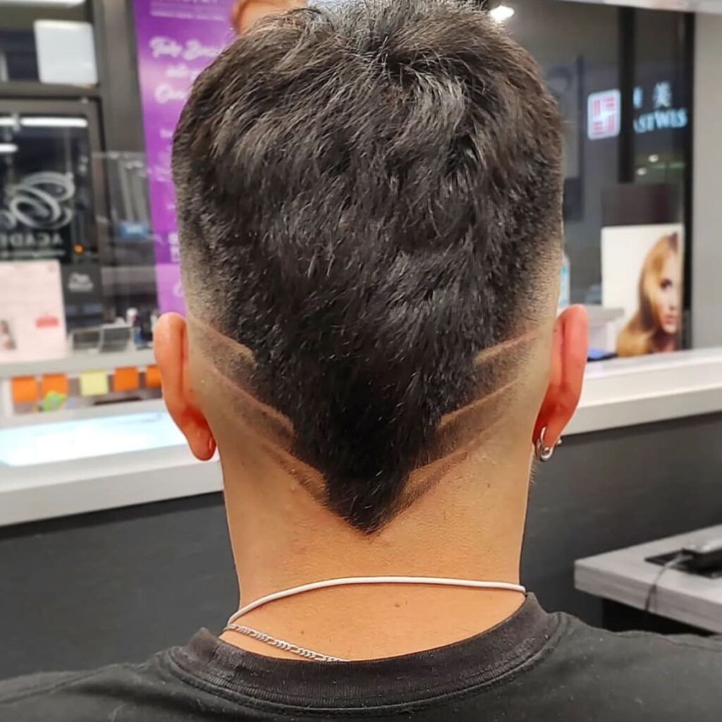 A back view of a man showcasing a bold and creative hairstyle with a precise geometric design shaved into the faded sides. The haircut features a sleek, tapered fade that blends seamlessly into the longer hair at the center, styled into a sharp V-shaped point at the nape of the neck. The clean, angular lines of the shaved design create a striking visual effect, adding a modern and personalized touch to the look. The overall style exudes confidence and individuality, perfect for someone who wants to make a statement. The image is taken in a barbershop setting, highlighting the artistry and detail of the hairstyle.