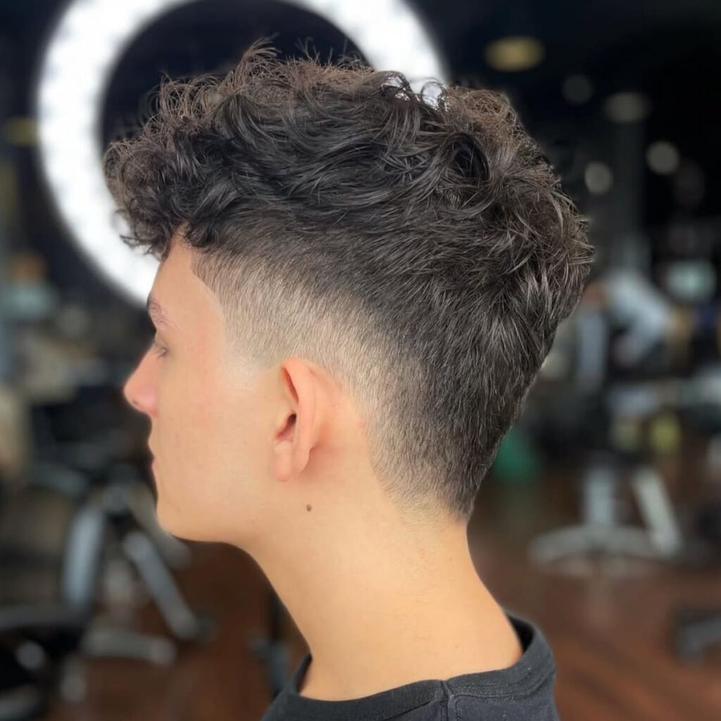 A side profile of a young man with a tapered haircut that gradually transitions from a clean, low fade on the sides to longer, curly hair on top. The curls are naturally styled, adding volume and texture, while the fade provides a sleek and polished look. The subtle taper at the neckline enhances the overall sharpness of the cut, blending seamlessly with the natural flow of the hair. The image is taken in a barbershop setting with a circular light in the background, drawing attention to the clean details of the hairstyle. The overall style combines modern sophistication with a casual, effortless vibe.