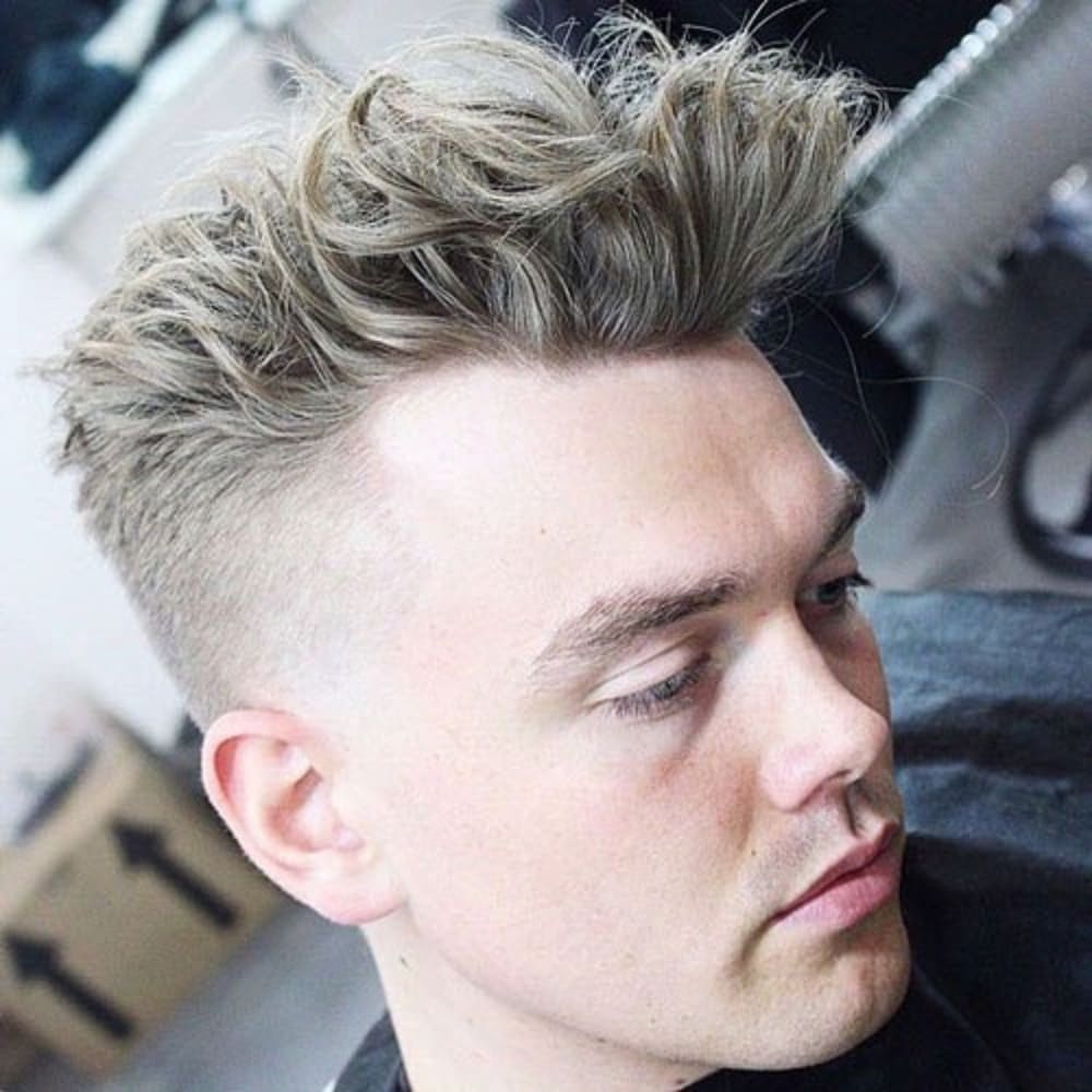 A close-up side profile of a man with voluminous, textured hair styled upwards and slightly tousled, featuring a clean fade on the sides. The hair on top is thick and full, with defined texture and a natural lift. The man has smooth, fair skin and a relaxed expression, with soft lighting enhancing the details of the hairstyle. The background is a neutral salon setting, providing a clean and professional atmosphere.