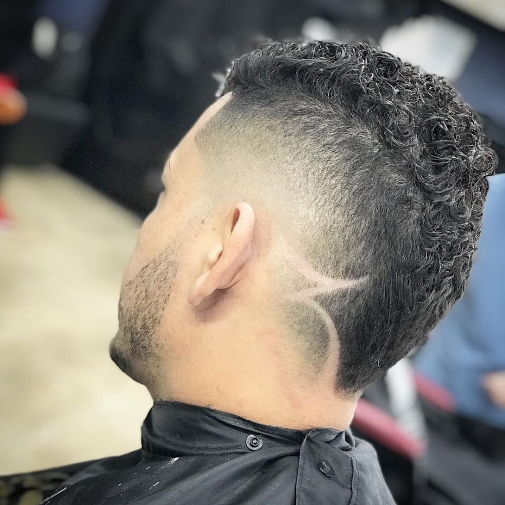 Curly mohawk fade hairstyle with sharp skin fade and intricate design shaved into the lower back of the head, showcasing bold texture and creative detail.