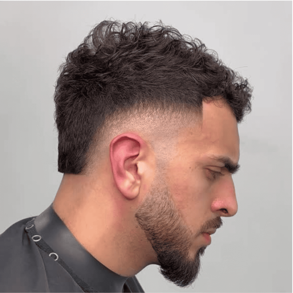 A side profile of a man with a clean, tapered haircut featuring textured curls on top blending seamlessly into the sides. The sharp lines of the cut enhance the neat, well-defined beard that frames his jawline. The contrast between the voluminous curls and the close taper creates a modern, polished look. The attention to detail in both the hair and beard adds to the overall sharp and stylish appearance