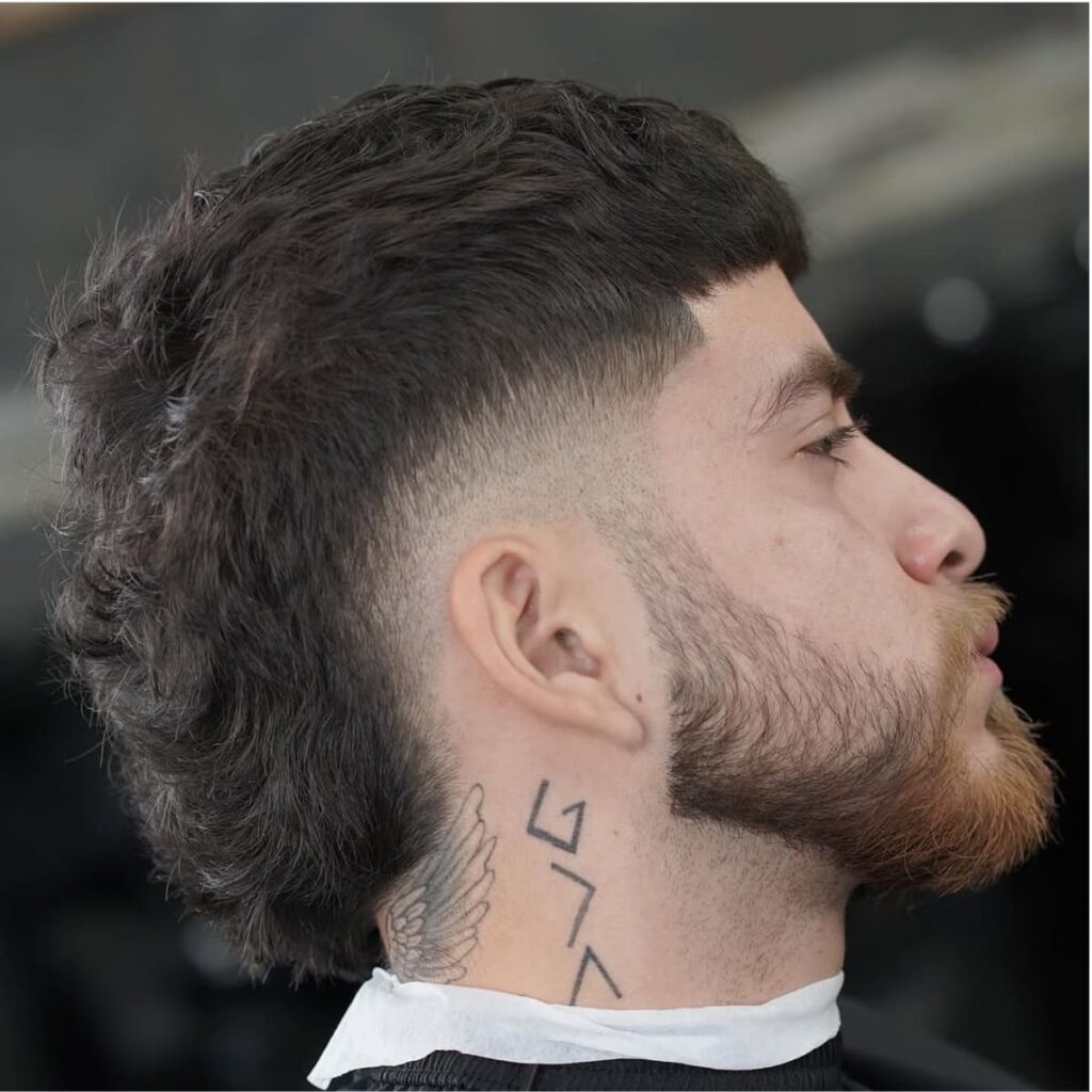 A side profile of a man with a bold bald burst fade haircut seamlessly transitioning into a modern mullet at the back. The fade curves sharply around the ears, creating a clean and defined contrast with the textured top and flowing mullet. The man’s beard is meticulously groomed, complementing the sharp lines of the fade and adding a rugged edge. Visible tattoos on the neck enhance the overall edgy and contemporary vibe of the look.