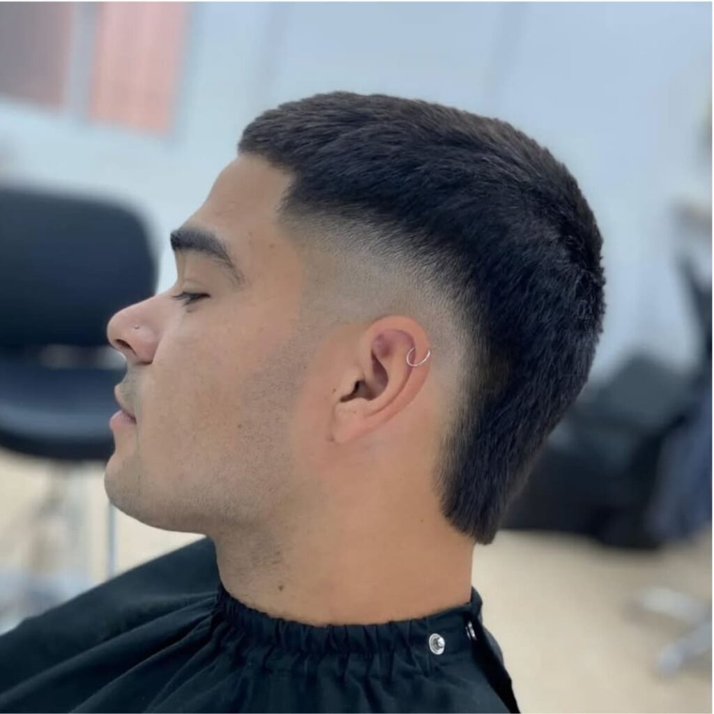 A side profile of a man showcasing a clean bald burst fade haircut with a buzzed top. The fade gradually blends into the skin around the ears, highlighting sharp, precise lines for a sleek and minimalist look. The neatly trimmed buzz complements the fade, creating a tidy and polished style. The subtle taper at the back enhances the overall clean appearance, while a small hoop earring adds a touch of individuality.