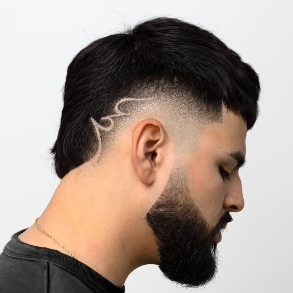 A side profile of a man featuring a clean and sharp bald burst fade haircut. The fade transitions smoothly into a creative shaved design along the side, adding a unique and artistic touch. The top is kept neat and textured, while the groomed beard complements the overall sharp and polished look. The intricate detail of the design enhances the modern and bold style.