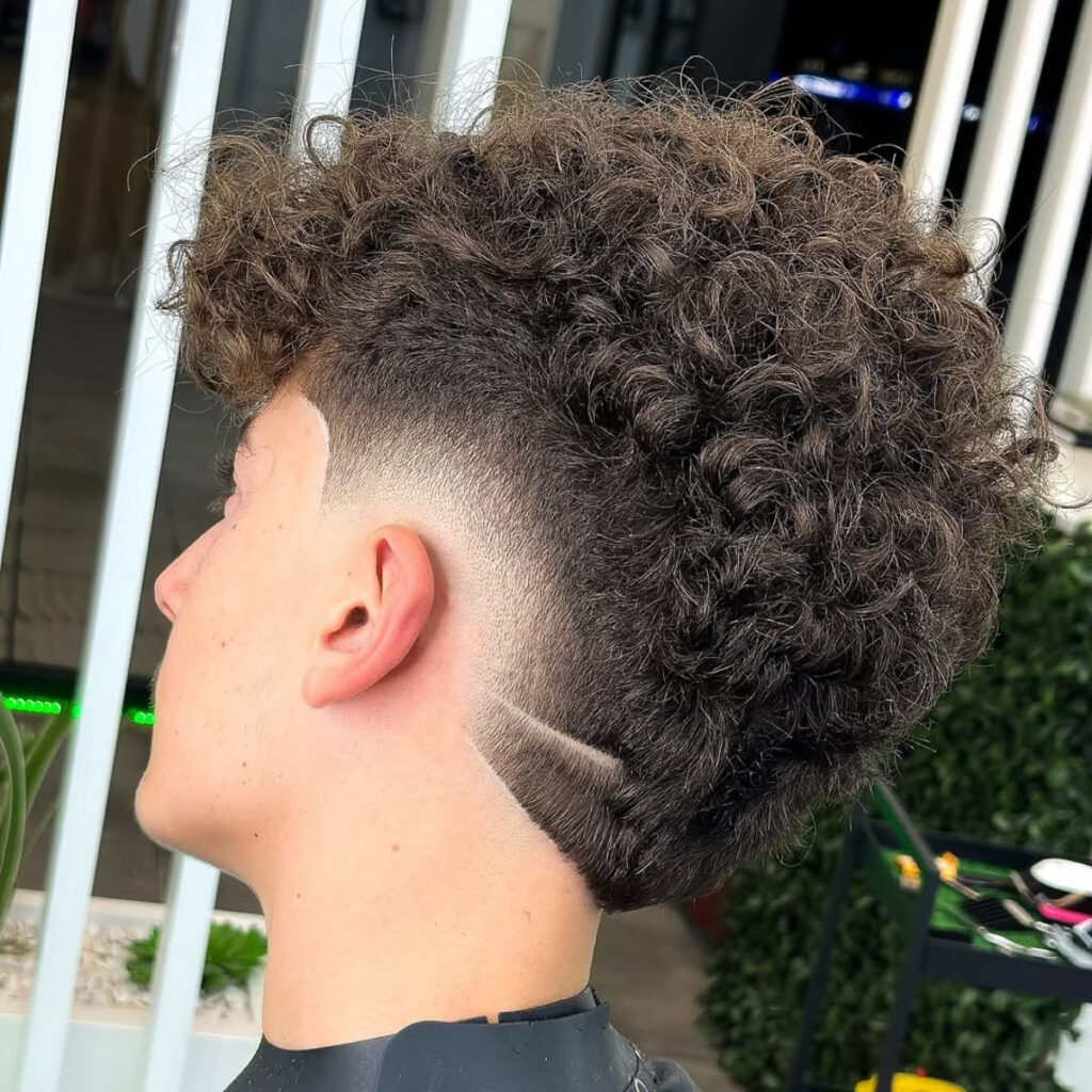 A side profile of a man with voluminous, curly hair on top, transitioning into a sharply tapered cut around the ears. The cut curves smoothly towards the nape of the neck, with a precise shaved line detail adding a stylish touch. The natural texture of the curls contrasts beautifully with the clean and sharp taper for a modern and dynamic look.