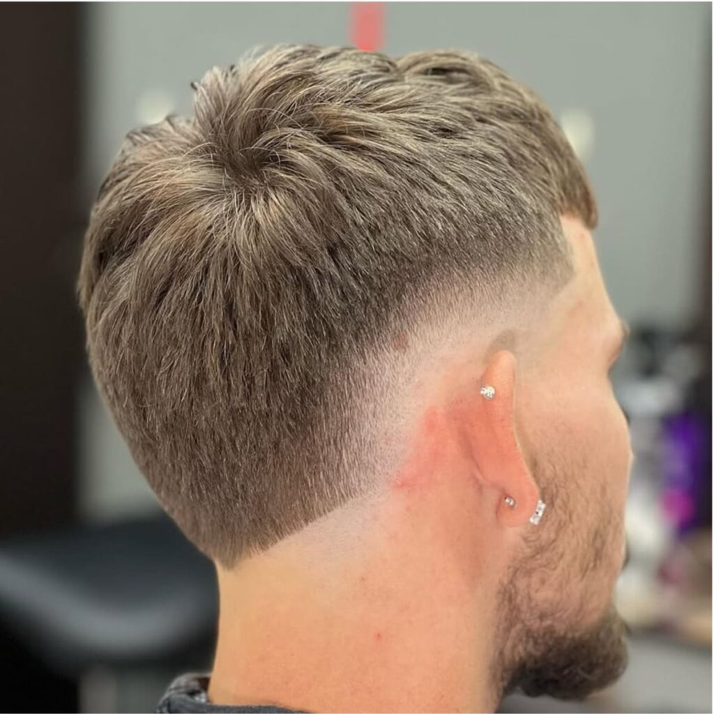 A rear-side profile of a man showcasing a clean bald burst fade haircut. The fade transitions smoothly from the skin near the ears to a longer, textured top, creating a sharp and balanced look. The haircut features precise blending with defined edges around the neckline for a polished finish. The natural texture on top adds depth and contrast to the sleek fade, highlighting the modern yet timeless appeal of the style. A subtle ear piercing adds a touch of personal flair.
