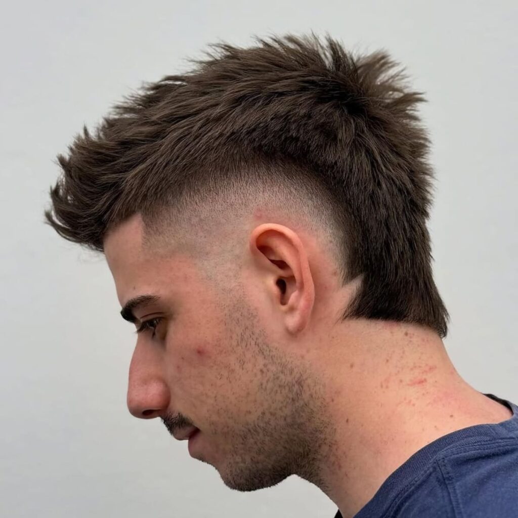 A side profile of a man sporting a high bald burst fade haircut. The fade curves sharply around the ears, transitioning into textured, voluminous hair on top for a bold and edgy look. The back features a modern mullet-style extension, adding a unique twist to the overall style. The clean fade contrasts perfectly with the rugged texture of the top and back, creating a trendy, standout appearance.