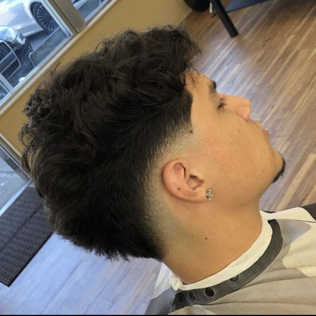 A side profile of a man with voluminous, curly hair on top that flows into a clean, tapered cut around the sides. The cut gradually blends towards the lower section near the ears and nape, giving a polished yet natural look. The texture on top contrasts beautifully with the smooth taper, creating a balanced and stylish appearance. The man’s sharp grooming and subtle goatee add to the overall modern vibe.