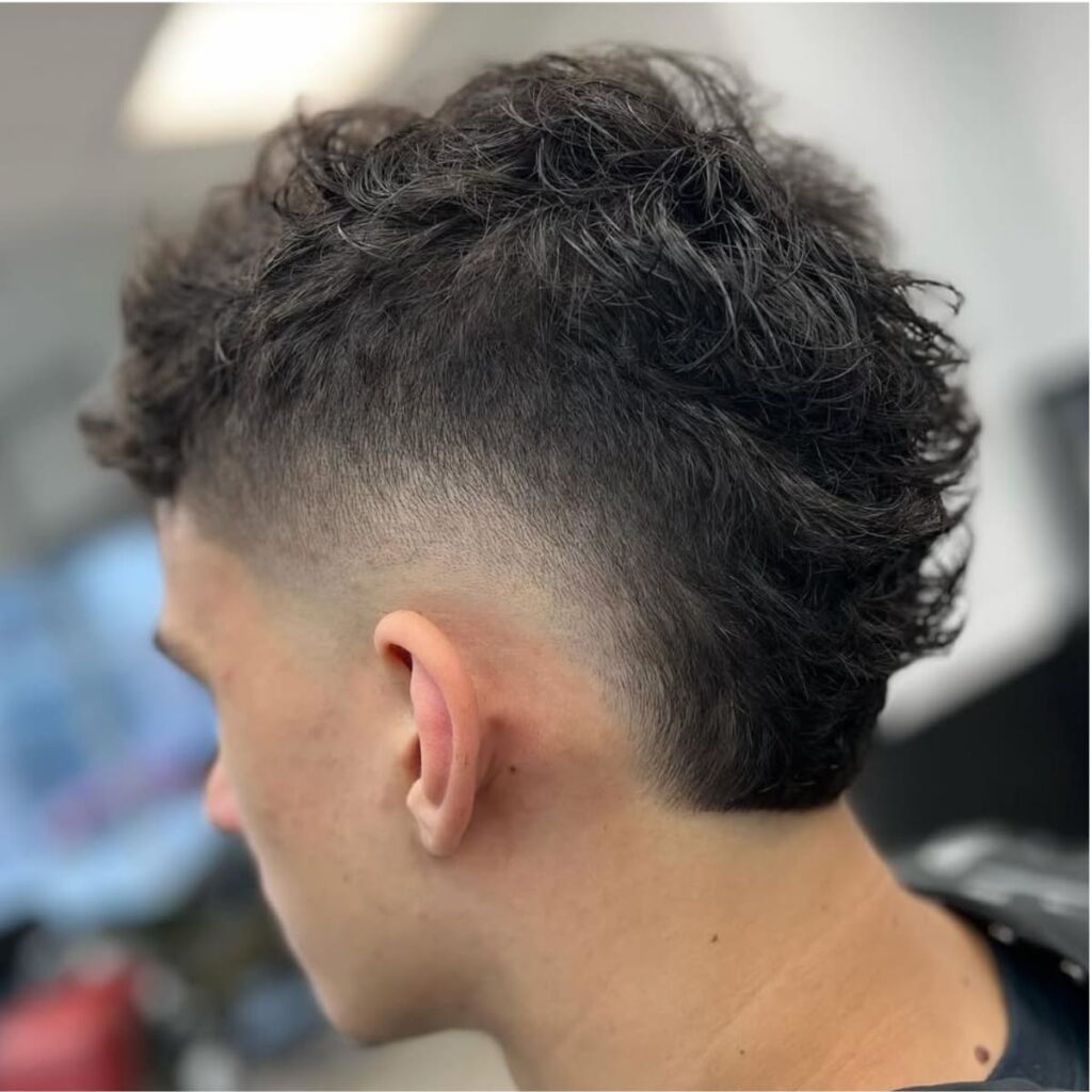 A back-side profile of a man with a textured bald burst fade haircut. The fade transitions smoothly from the skin around the ears into a voluminous, wavy top for a balanced and modern look. The haircut features a clean, rounded curve at the sides, adding sharpness to the style. The natural waves on top enhance the contrast between the clean fade and textured hair, making it a bold yet sophisticated choice.