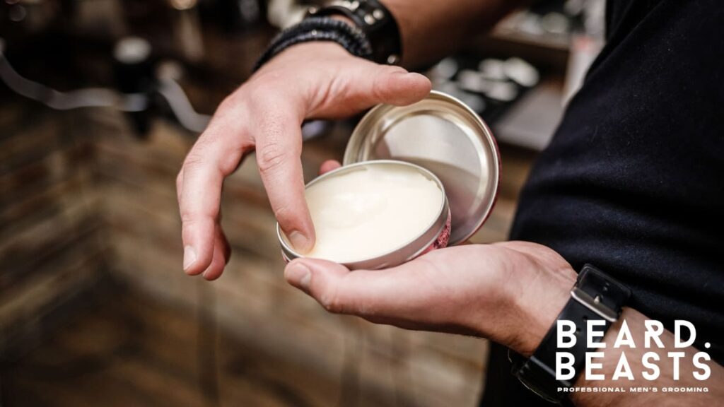 Close-up of a man scooping beard balm from a tin, showcasing the use of grooming products for styling and maintaining a healthy, well-groomed curly beard.