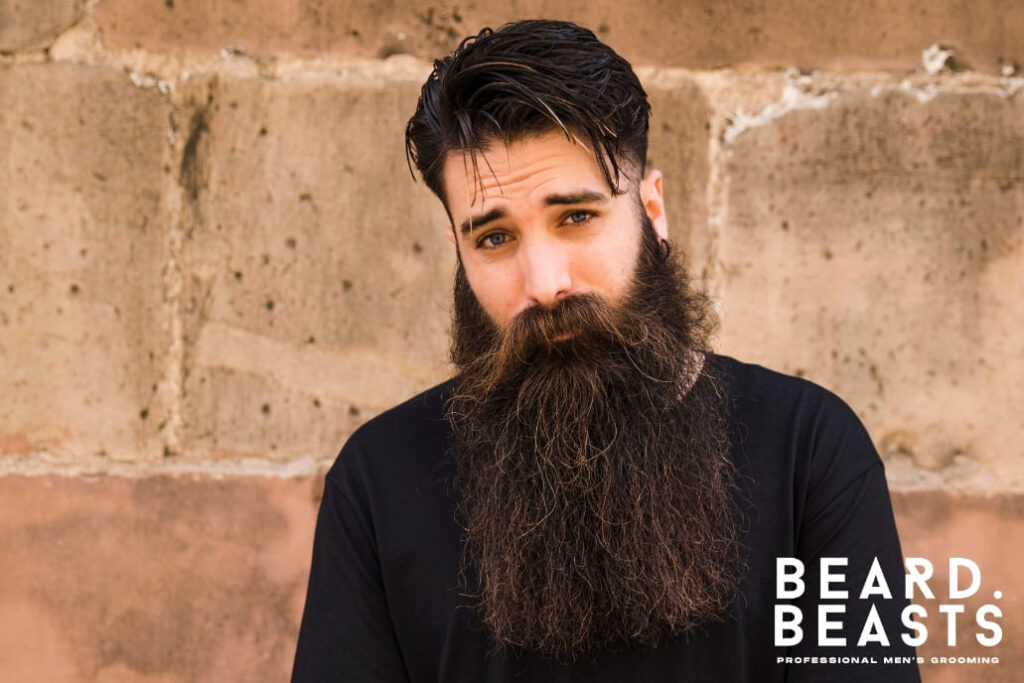 Man with a healthy, long, and well-detangled beard standing against a rustic wall, showcasing proper beard care and grooming techniques for learning how to detangle a beard.