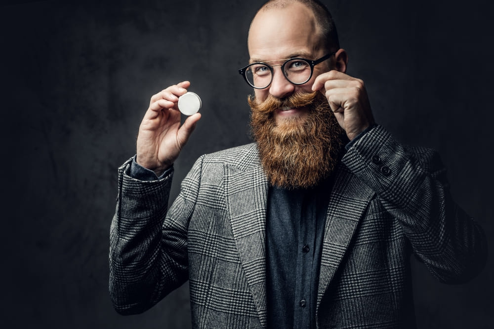 A bald man with a full beard and styled mustache holding a tin of mustache wax while shaping his mustache, demonstrating proper grooming techniques.