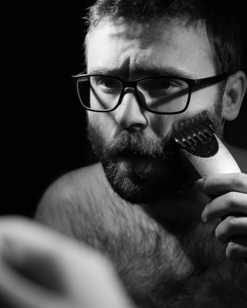 Man trimming his beard in the mirror, showing how the right beard shape and length can make facial hair look thicker without exposing weak areas.