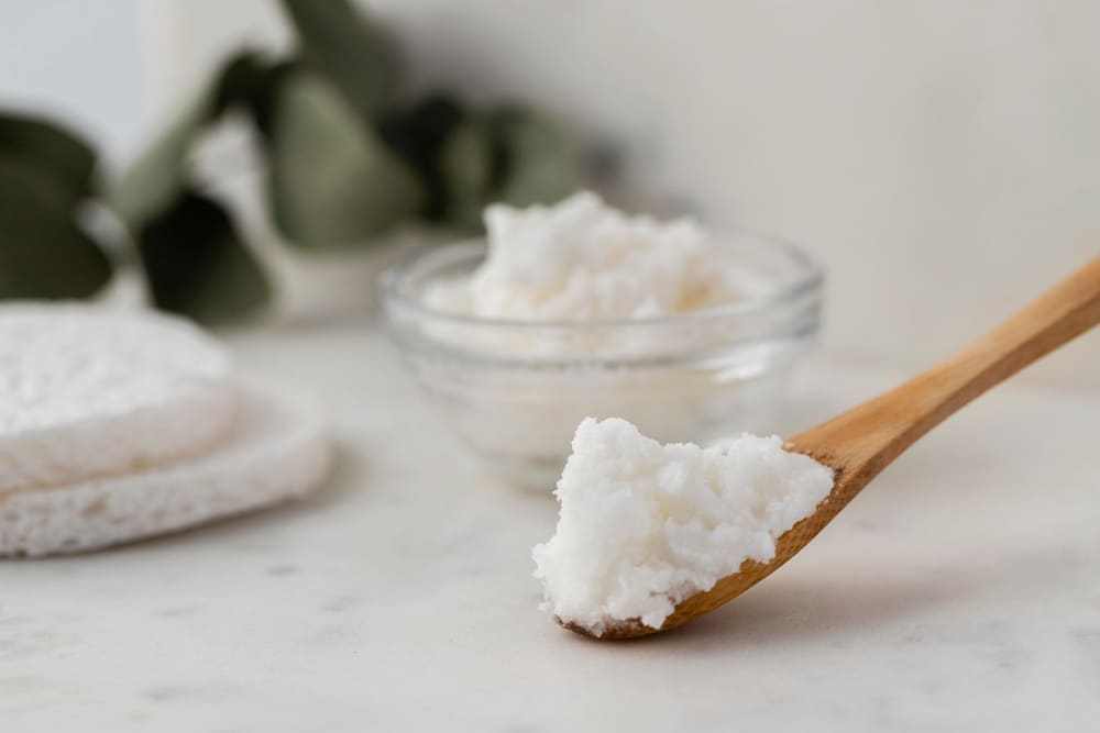 Wooden spoon holding a creamy white substance, placed on a marble surface with a glass bowl of the same substance and soft pads in the background, suggesting a natural and soothing product for skin or hair care.