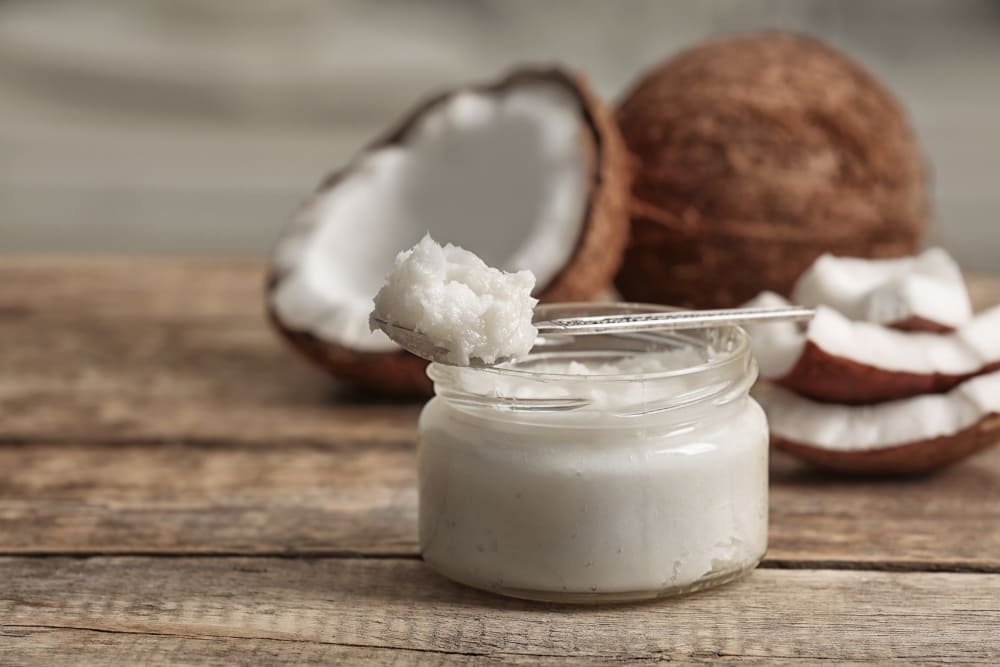 Jar of coconut butter with a spoonful resting on top, surrounded by fresh coconut halves and slices on a rustic wooden table, emphasizing its natural use in beard care.