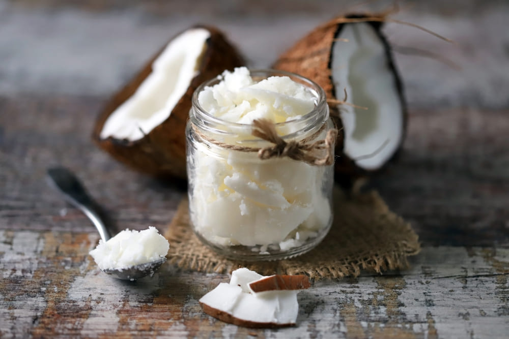 Glass jar of coconut butter tied with rustic twine, surrounded by fresh coconut halves, coconut pieces, and a spoon on a wooden surface, highlighting its use in coconut butter for beard care.