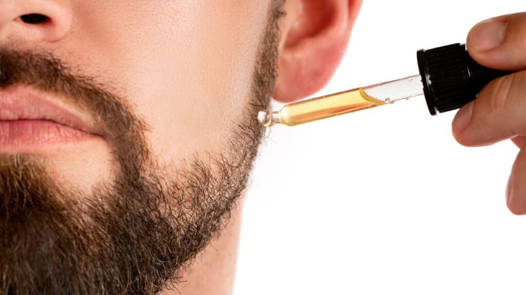 A close-up of a man applying beard oil with a dropper to his beard, emphasizing the importance of hydration and nourishment.