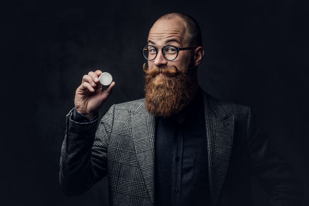 A bearded man with a styled mustache holding a tin of mustache wax, showcasing the essential product for achieving a well-groomed look. A perfect visual for tutorials on how to apply mustache wax effectively.