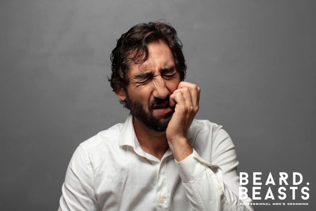 Man in a white shirt scratching his beard due to discomfort caused by beard rash, set against a neutral gray background. Beard Beasts logo is visible in the bottom-right corner, emphasizing professional men's grooming.