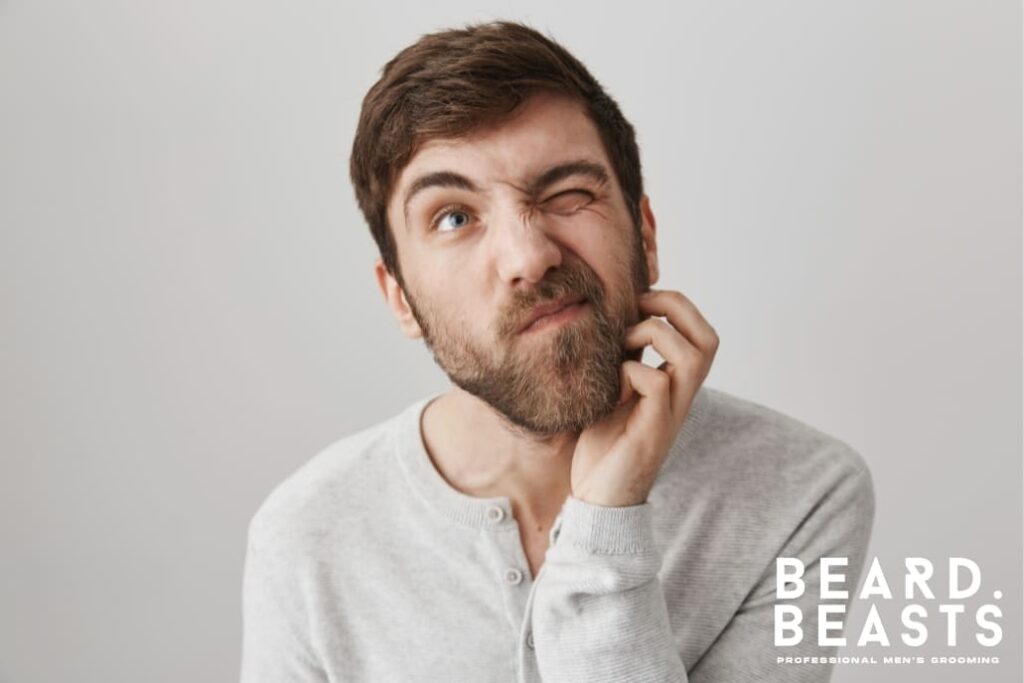 Young man in a light gray shirt scratching his beard while grimacing in discomfort, likely due to beard rash. The neutral background highlights the subject, and the Beard Beasts logo in the bottom-right corner emphasizes professional men's grooming.