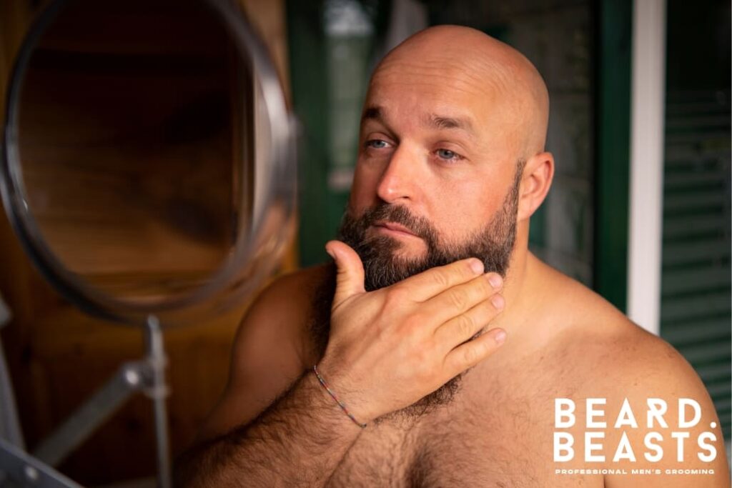 Man with a full beard in a light gray shirt scratching his facial hair with a thoughtful expression. The image features a neutral gray background and includes the Beard Beasts logo in the bottom-right corner, symbolizing professional men's grooming.