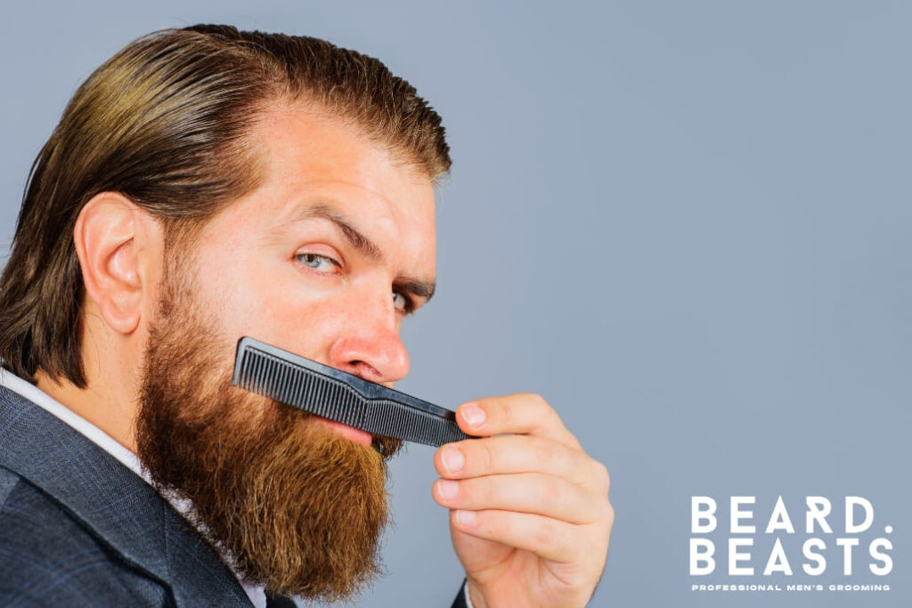 A professional man with a well-groomed beard combing it using a plastic comb, wearing a suit, and posing confidently against a neutral gray background.